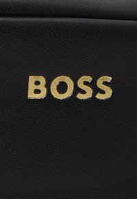 Melnas ādas materiāls ar struktūrētu virsmu, kurā izceltas vārds "BOSS" treknām, reljefām zeltītām burtiem.
