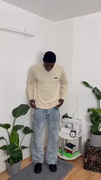 Suéter bege de manga longa com um pequeno logo, combinado com jeans rasgados de tom azul claro. Sapatos pretos. Ambiente interno com plantas e móveis.