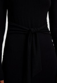 Vestito nero a coste con maniche lunghe, dotato di un dettaglio in vita legato. Il tessuto sembra morbido ed elastico, creando una silhouette aderente.