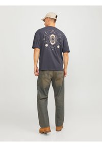 Dunkelgraues T-Shirt mit großem himmlischen Grafikdesign auf dem Rücken, kombiniert mit lockeren, ausgebleichten grünen Jeans und brauen Schuhen.
