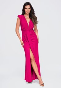 Femme aux longs cheveux foncés portant une robe sans manches rose foncé avec un décolleté en V profond et une fente haute à la cuisse, posant sur un fond clair.