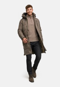 Zimná bunda s kapucňou v olivovo zelenej farbe, so zipsom vpredu a bočnými vreckami, nosená cez svetlohnedý sveter a tmavé džínsy. Hnedé šnurovacie topánky.