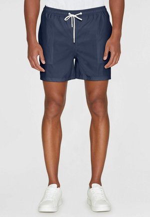 Mand iført marineblå badeshorts med hvid snor og hvide sneakers, stående mod en ensfarvet baggrund.