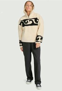 Κρεμ pullover με μαύρο αφηρημένο σχέδιο, κλείσιμο με φερμουάρ στον λαιμό και φαρδιά μανίκια, σε συνδυασμό με σκούρους παντελόνες και μαύρα αθλητικά παπούτσια.