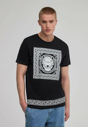 T-shirt in cotone nero con una grafica di leone bianco all'interno di un bordo decorativo, con un motivo a chiave greca lungo l'orlo.