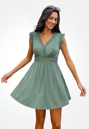 Vrouw draagt een mouwloze groene eyelet jurk met gegolfde V-hals en kanten taille, glimlachend met een zonnebril op haar hoofd voor een effen achtergrond.