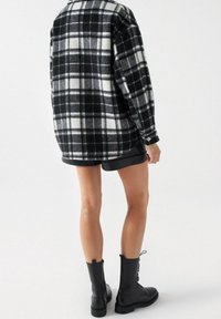 Svart och vit rutigt flanellskjorta med långa ärmar, bärs över svarta shorts. Modellerna har svarta snörboots synliga.