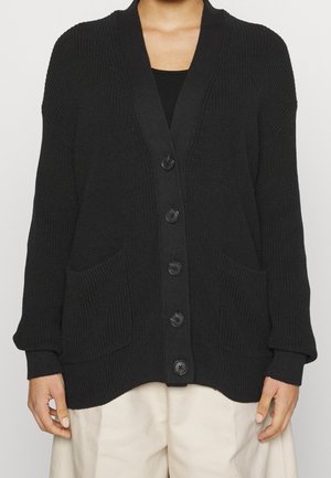 Zwarte gebreide cardigan met knopen en twee voorzakken, gedragen over een zwart topje en beige broek.