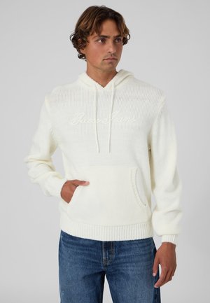 Homme portant un sweat à capuche blanc en tricot avec une poche avant et un jean bleu, debout devant un fond uni gris clair.