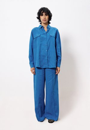 Chemise oversize bleue avec deux poches poitrine, assortie à un pantalon large. Tissu lisse avec une finition mate, arborant un col.