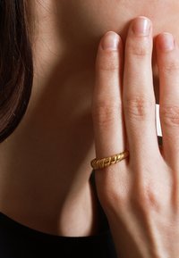 Goldene, verdrehte Bandring, der an einem Finger getragen wird und ein strukturiertes Design mit glatten und geriffelten Abschnitten auf nackter Haut zeigt.
