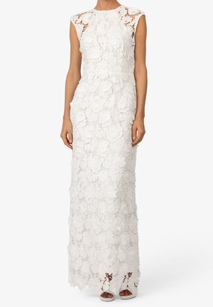 Robe blanche sans manches longue jusqu'au sol avec superposition en dentelle florale et col montant, portée avec des talons ouverts blancs.