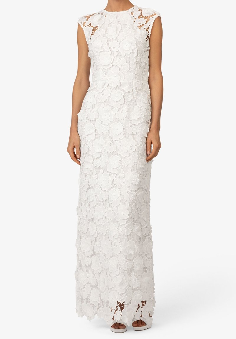 Robe blanche sans manches longue jusqu'au sol avec superposition en dentelle florale et col montant, portée avec des talons ouverts blancs.