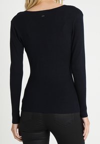 Mörkblå ribbad stickad tröja med långa ärmar, bred rund halsringning och smal midja. Slät textur med subtilt varumärkeslogotyp på baksidan.