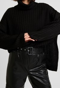 Pull en maille noire à col roulé avec des manches oversize, associé à un pantalon en cuir noir taille haute avec une large ceinture et un détail de boucle.