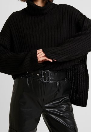 Zwarte geribbelde coltrui met oversized mouwen, gecombineerd met zwarte leren high-waisted broek met een brede riem en gesp-detail.