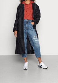 Abrigo negro a rayas, sudadera roja y vaqueros azules desgastados. Zapatillas blancas y marinas. Enfocado en los patrones del tejido y el corte relajado.
