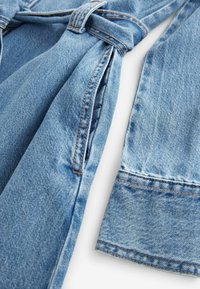 Manche et détail de poche d'une veste en denim bleu clair avec coutures visibles et passants de ceinture sur fond blanc.