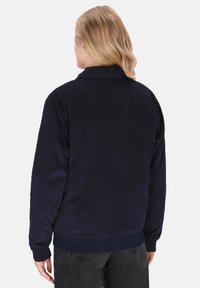 Persoon met blond haar, gekleed in een marineblauwe corduroyjas en zwarte jeans, staande met de rug naar de camera tegen een effen achtergrond.