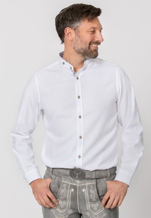 Smiling man wearing a white buttoned shirt and traditional gray embroidered lederhosen with a stag belt buckle, hands in pockets.