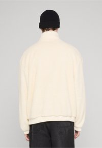 Cream-colored fleece jacket with a high collar, drop shoulders, and ribbed cuffs. Worn with a black knitted beanie. Soft texture.