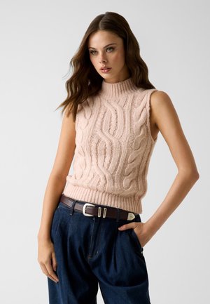 Frau mit langem braunem Haar trägt einen ärmellosen pinkfarbenen, kabelgestrickten Pullover und hoch taillierte dunkelblaue Jeans mit braunem Gürtel, steht mit einer Hand in der Tasche.