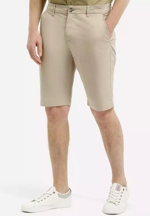 Beżowe bawełniane szorty z płaskim przodem, kieszeniami bocznymi i zapięciem na guzik. Ubrane z białymi tenisówkami z szarymi akcentami.