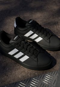 Baskets Adidas noires avec des rayures blanches et des lacets posées sur une surface texturée avec un éclairage ombré.