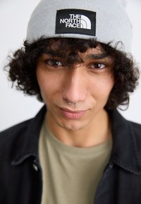 Beanie grigia a maglia con patch logo nero, indossata su abbigliamento scuro. Capelli ricci scuri e piercings sul viso. Caratteristiche facciali ben definite su uno sfondo chiaro.