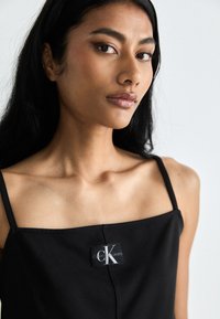 Svart ermeløs topp med firkantet hals, med en sydd Calvin Klein Jeans-merkelapp i midten. Glatt stoff og minimalistisk design.
