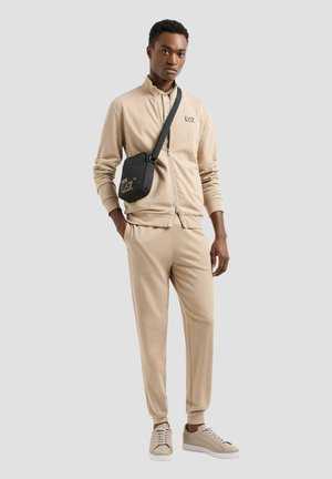 Giovane uomo con tuta beige e sneakers con borsa nera a tracolla, in piedi davanti a uno sfondo grigio chiaro uniforme.