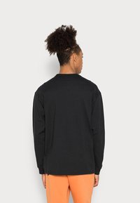 Μαύρο μακρυμάνικο μπλουζάκι με χαλαρή εφαρμογή, στρογγυλή λαιμόκοψη και λοξές μανσέτες; λείο βαμβακερό ύφασμα; φορεμένο με πορτοκαλί παντελόνια.