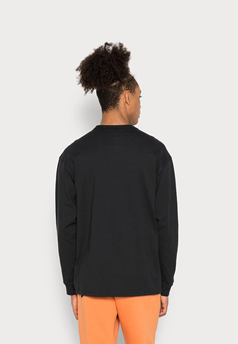 Schwarzes langärmliges T-Shirt mit lockerer Passform, Rundhalsausschnitt und gerippten Bündchen; glattes Baumwollmaterial; getragen mit orangefarbenen Hosen.