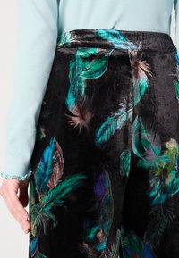 Pantaloni din catifea cu un fundal negru și imprimeuri cu pene multicolore în verde, albastru și portocaliu. Prezintă o textură netedă și un talie ascunsă.