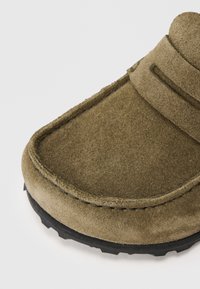 Olivgrüner Wildlederschuh zum Hineinschlüpfen mit runder Spitze, kontrastierender schwarzer Gummisohle und genähten Akzenten auf dem Obermaterial.