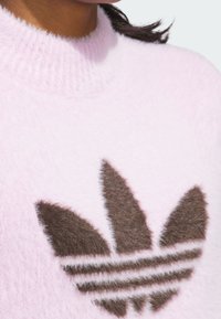 Lyserød, fluffy sweater med ribbet halsudskæring, der har et brunt, fluffy logo-design og tre vandrette striber nederst på logoet.