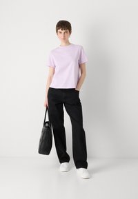 Camiseta corta de manga corta en color púrpura claro con cuello redondo, combinada con pantalones anchos negros y zapatillas blancas. Sostiene un bolso texturizado negro.