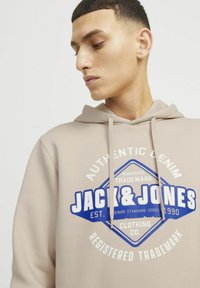 Beige hoodie med snoretræk og frontgrafik, der viser "JACK&JONES" i blå og hvid. Blød stoftekstur med en afslappet pasform.