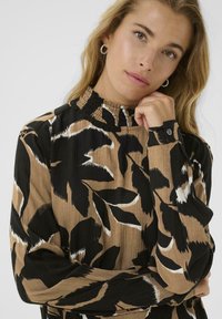 Vrouw met blond haar, draagt gouden ringoorbellen en een bruin met zwart bladpatroon lange mouw shirt, leunend met haar kin op haar hand.