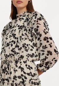 Svart blommönster på genomskinlig krämfärgad tyg, med volanger vid axlar och bröst, elastisk midja och mjuk känsla.