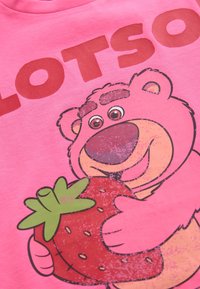 Ružové tričko s obrázkom kresleného medveďa držiaceho jahodu, nad ktorým je červený nápis "LOTSO". Tričko má hladkú textúru.