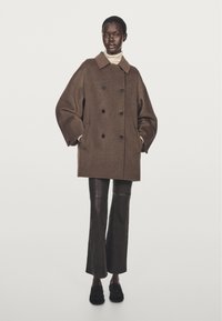 Brun ullkappa med dubbelknäppt design, stor krage och svarta knappar, kombinerad med svarta läderbyxor med utsvängd fot och mörka skor.