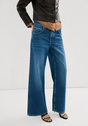 Modelo con jeans anchos azules, camisa marrón a rayas, chaqueta de cuero negra y sandalias negras con tacón.