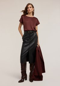 Kvinna klädd i en bourgognefärgad satinblus, svart läder midi-kjol med slits fram, bruna ormskinnsstövlar och håller en matchande bourgognefärgad blazer.