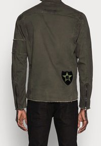 Olivengroene militaire jas met een onafgewerkte zoom, ritsen op de mouwen en een zwart patch met een ster op de achterkant.