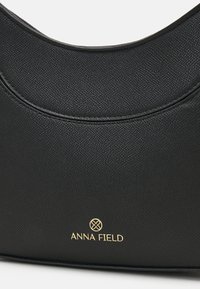 Musta tekstuuripintainen nahkalaukku, jossa on kaareva yläreuna ja kultainen ANNA FIELD -logo ja emblem, joka sijaitsee keskellä alaosaa.