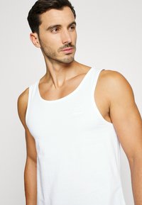 Débardeur blanc en coton, avec un col rond et un petit logo sur la poitrine. Texture lisse sans motifs ni accents.