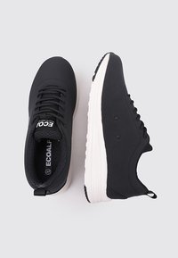 Zapatillas negras con un upper de tejido suave, forma redondeada, suela de goma blanca y cordones planos; presenta una marca en la lengüeta.