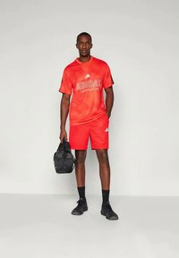 Conjunto desportivo vermelho da Adidas, com uma t-shirt de manga curta com logótipo e calções a condizer. Inclui uma mala de ginásio preta e sapatilhas atléticas pretas.