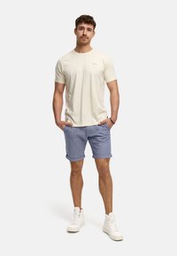 Helles beige Kurzarmhemd mit dezentem Logo, kombiniert mit hellblauen Shorts, aufgerollten Manschetten und weißen hochgeschnittenen Sneakers.